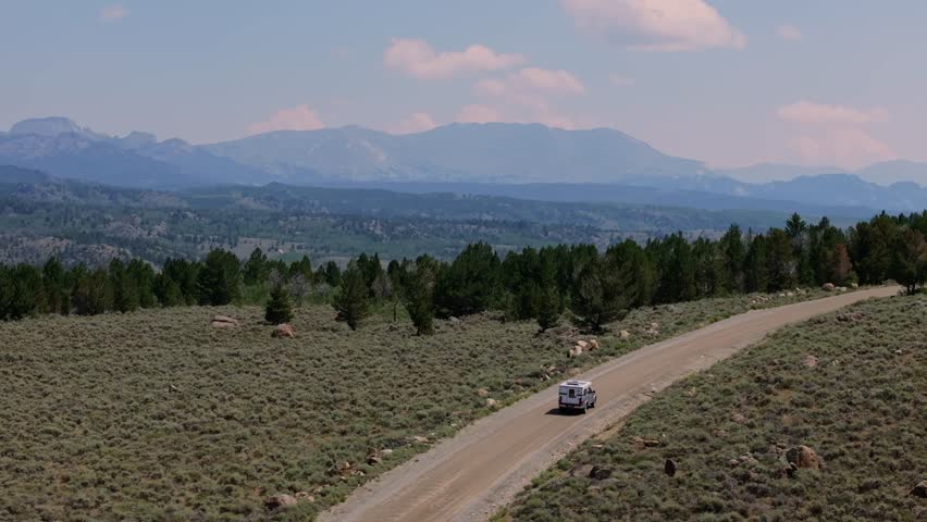 Aerial rearview orbit follows camper truck driving on dirt road in Wyoming’s Wind River Range, showcasing wide plains and mountains