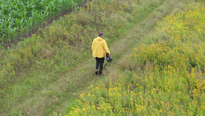 A man in a yellow raincoat and umbrella strolls on a narrow path amid fields and trees in the rain. Outdoor relaxation and physical activity. Filmed in slow motion.