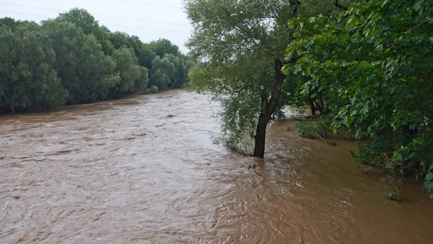 A flooded river seen from a bridge after torrential rain, water covering trees and fields. Debris-laden water churns, recorded in slow motion. Natural disaster.