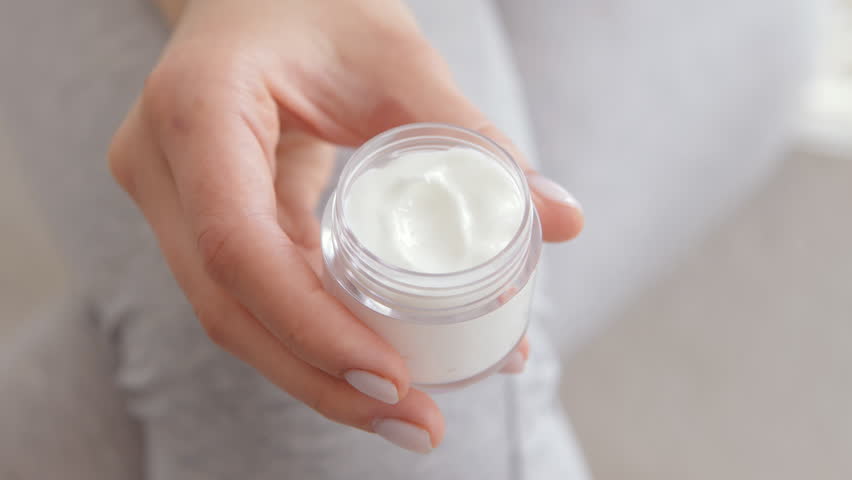 A hand grips a clear jar containing white cream, set against a soft, bright background. The focus is on the moisturizer, highlighting its texture and freshness.