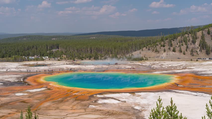 A vibrant timelapse capturing tourists walking around the iconic Grand Prismatic Spring in Yellowstone, with colorful geothermal activity and steam rising into the bright sky.