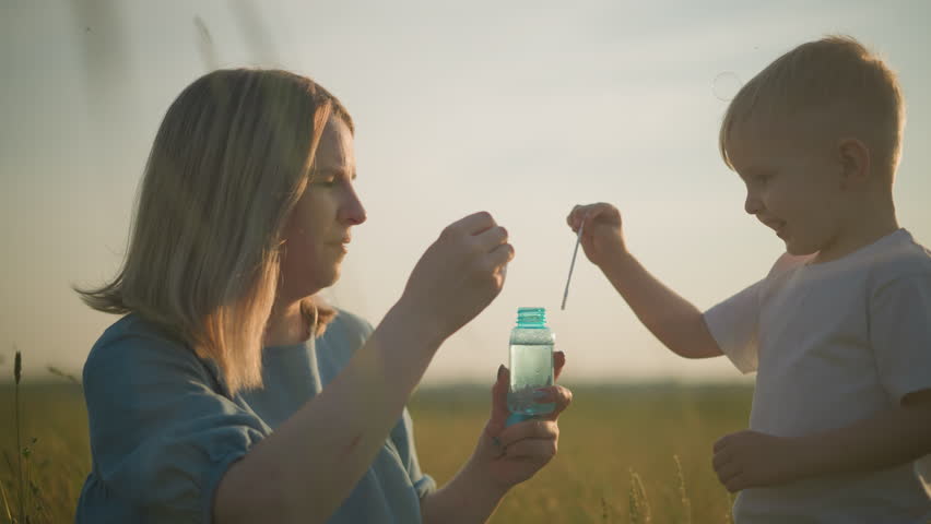 A close-up of a woman in a blue dress kneeling in a grassy field at sunset, holding a bubble bottle as she and her young son, dressed in a white shirt, blow bubbles from wands