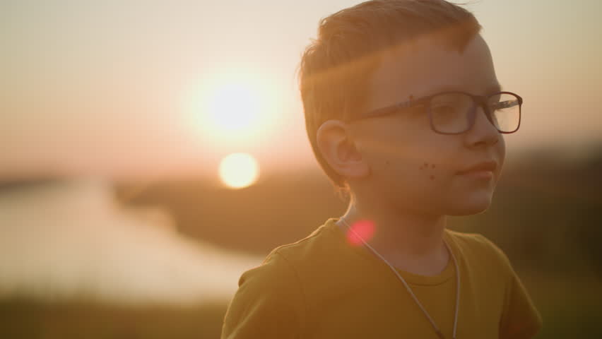Close-up of a young boy wearing glasses and a yellow shirt, smiling shyly while playfully moving his hands in the soft, warm glow of the sunset. His gentle expression is a moment of joyful innocence