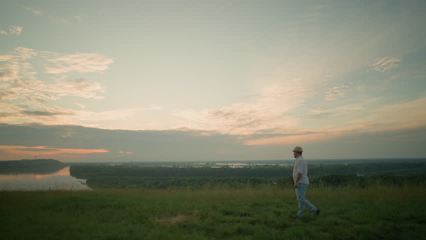 A man dressed in a white shirt, hat, and jeans walks through a grassy field towards a serene lake at sunset. He pauses with his arms folded, contemplating the peaceful landscape and tranquil waters