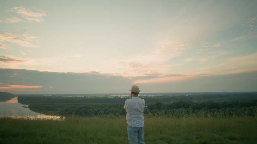A thoughtful man wearing a white shirt, hat, and jeans stands in a grassy field by a tranquil lake at sunset, gazing into the distance. The warm sunset light create a peaceful atmosphere