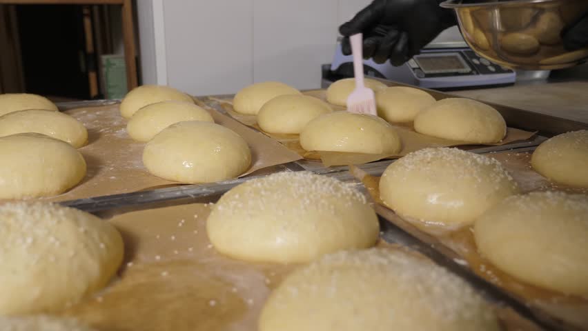 Close-up of the chef using a brush to apply egg mixture to round burger buns in the restaurant kitchen. The process of making dough for burgers.