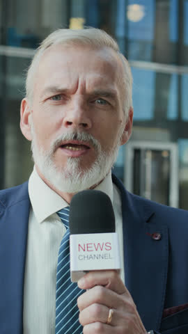 Vertical chest up of Caucasian mid-aged male reporter in blue suit and striped tie holding microphone while covering news for TV report, standing in front of glass office building in city downtown