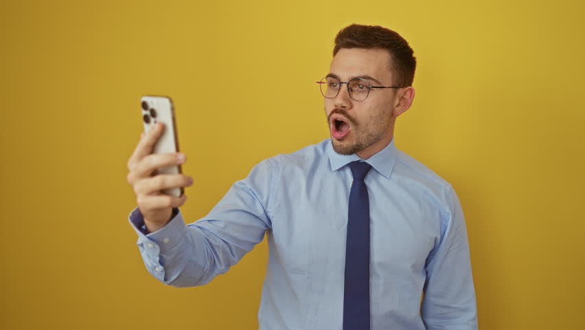 Young hispanic man in shirt and tie making a fish face with crazy eyes and mouth while using smartphone, over isolated yellow background, comical and silly expression