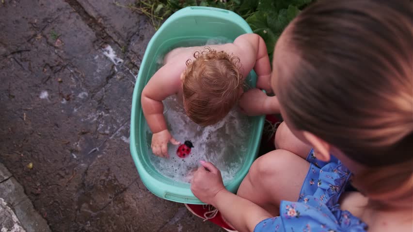 A joyful toddler enjoys bath time in a green tub while being lovingly supervised by a caring adult in a backyard setting