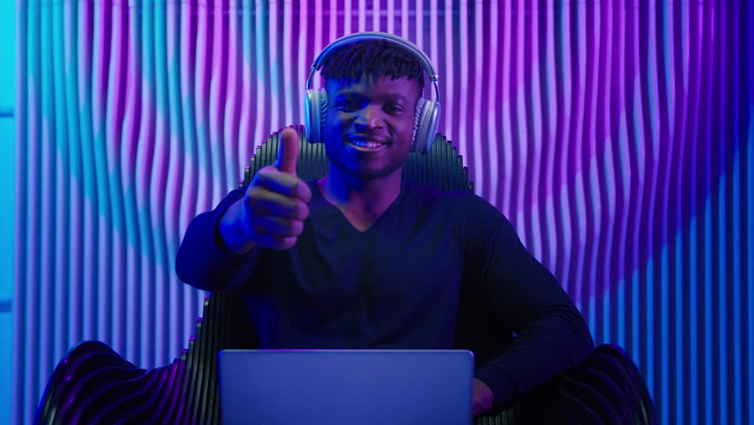 African American man guy male coder gamer programmer in chair wear headphones using laptop online work crypto software play internet game modern technology neon futurism showing thumb up like gesture