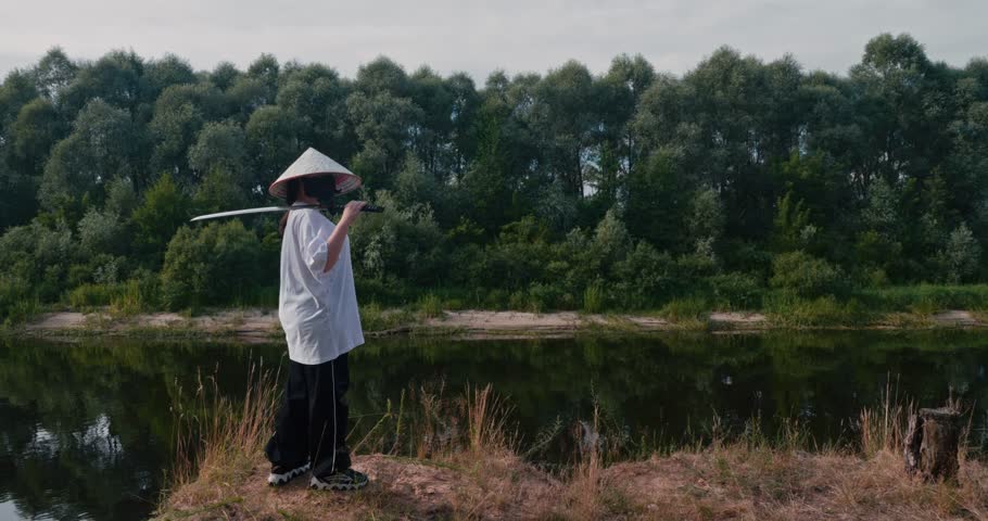 A beautiful woman on the edge of a cliff by the river with a katana in a mask and a straw hat. A samurai woman with a thoughtful look stands with a katana on a cliff and looks into the distance.