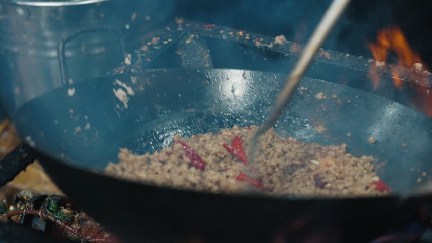 Anonymous person stirring minced beef with red pepper slices bubbling in oil in overheated wok pan over flame. Ground beef being browned in skillet with chopped vegetables during cooking process.