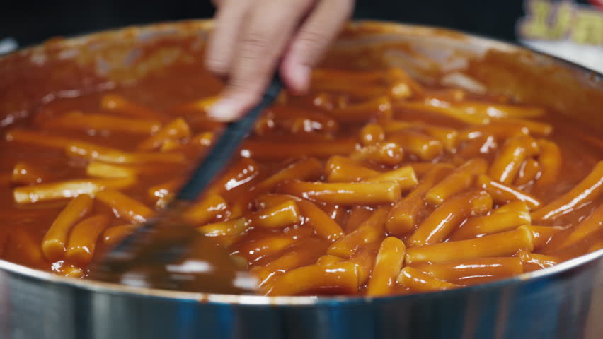 Full giant pan of tteokbokki simmered rice cake getting stewed in sauce made of red pepper paste and stirred with metal scoop. Faceless person preparing cylinder shaped noodle of korean cuisine.