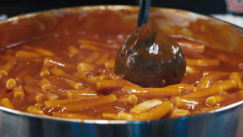Full giant pan of tteokbokki simmered rice cake getting stewed in sauce made of red pepper paste and stirred with metal scoop. Faceless person preparing cylinder shaped noodle of korean cuisine.