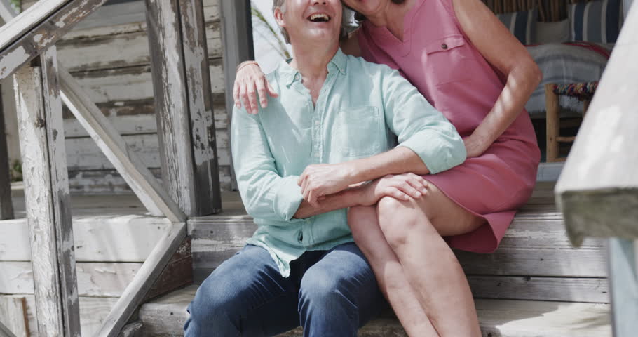 Portrait of happy senior caucasian couple embracing sitting on steps of beach house, in slow motion. Retirement, romance, relaxation, vacations and senior lifestyle.