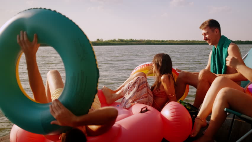 Group of friends in swimsuit having fun sitting on a pier toasting with beer, enjoying a summer day at the lake.