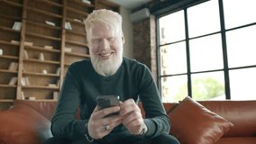 Young smiling albino man holding smartphone device texting sms message sitting at home office. Positive guy using apps playing mobile game chatting in social media surfing web on phone in modern home - Powered by Shutterstock - Get 15% off with code: PIKWIZARD15