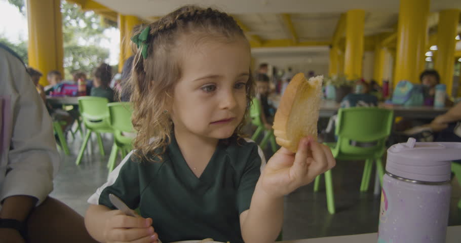 A blonde girl playfully enjoys a piece of bread in a cafeteria, her funny expressions and joyful energy capturing a fun and carefree breakfast moment.