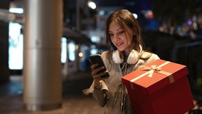 Beautiful Girl Walks Down the Street, Received New Year Greetings on Her Phone and Joyfully Reads It While Holding a Large Gift in Her Hands. Young Lady Outside Carries a New Year Gift and Uses Phone - Powered by Shutterstock - Get 15% off with code: PIKWIZARD15