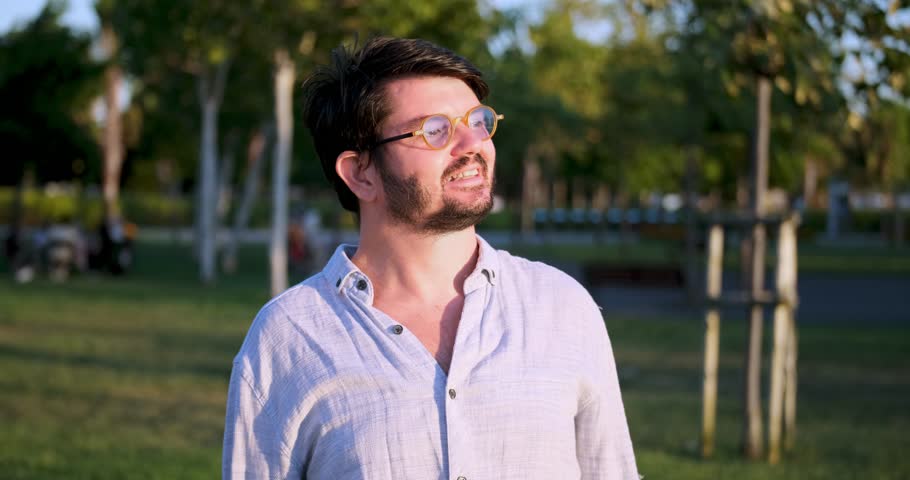 A middle-aged, dark-haired businessman wearing a shirt is smiling at the camera in a sunny, tree-filled park on a summer day