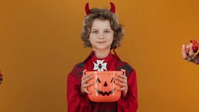 Medium shot of cheerful young blond Caucasian boy in fancy devil outfit, with horns holding Halloween pumpkin bucket for trick or treating, hands of people throwing candy, on orange background - Powered by Shutterstock - Get 15% off with code: PIKWIZARD15