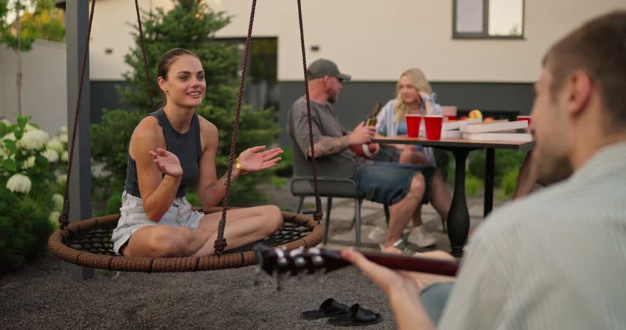 Over the shoulder a happy brunette girl claps and sings songs to the melody played by her boyfriend on the guitar during a party with his company in the backyard of a country house