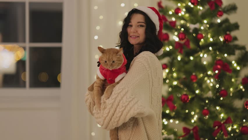A woman in a Santa hat and cozy sweater reacts in surprise while holding a ginger cat dressed in a Christmas outfit. The festive background features a beautifully decorated Christmas tree. Ideal for