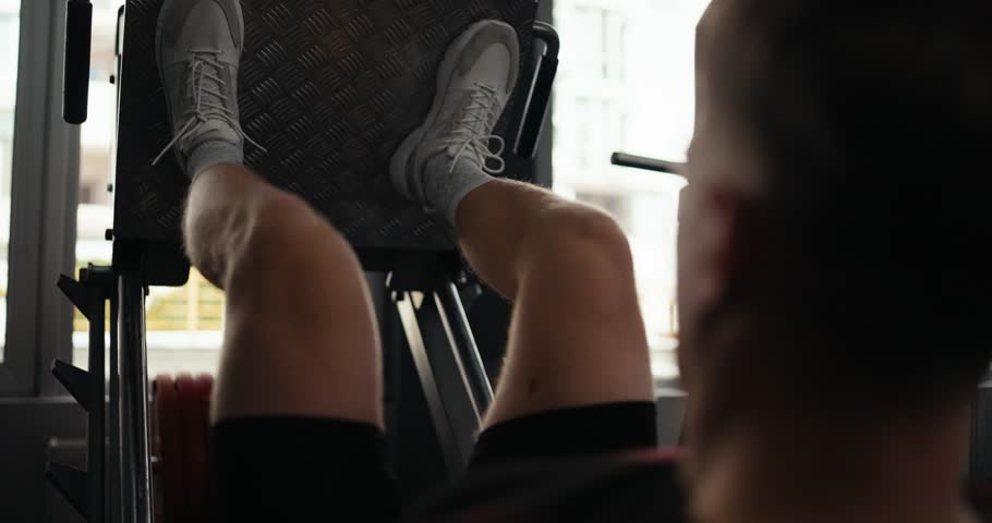 Over the shoulder up close a blond guy in gray sneakers performs a bench press during his leg workout in the fitness room
