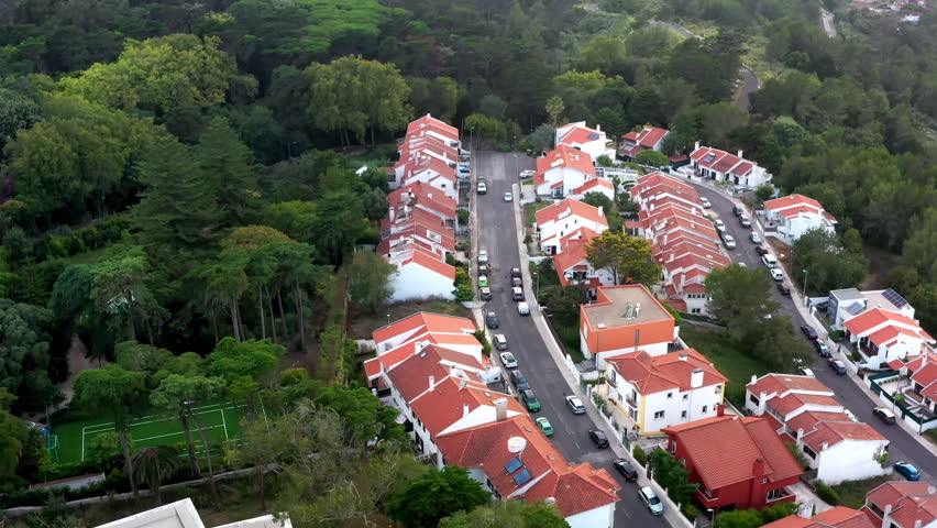 Drone footage suburban area in Sintra Portugal showcasing one-story and two-story homes near lush forest. Sports field is visible, highlighting area's community vibe and scenic surroundings