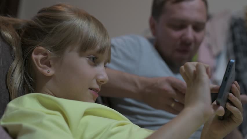 Dad and daughter sit at home and play video games on a mobile phone