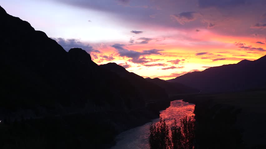 This landscape photo captures a stunning sunset over a calm river. The sky glows orange and pink, while lush mountains and a serene meadow complete the peaceful scene.
