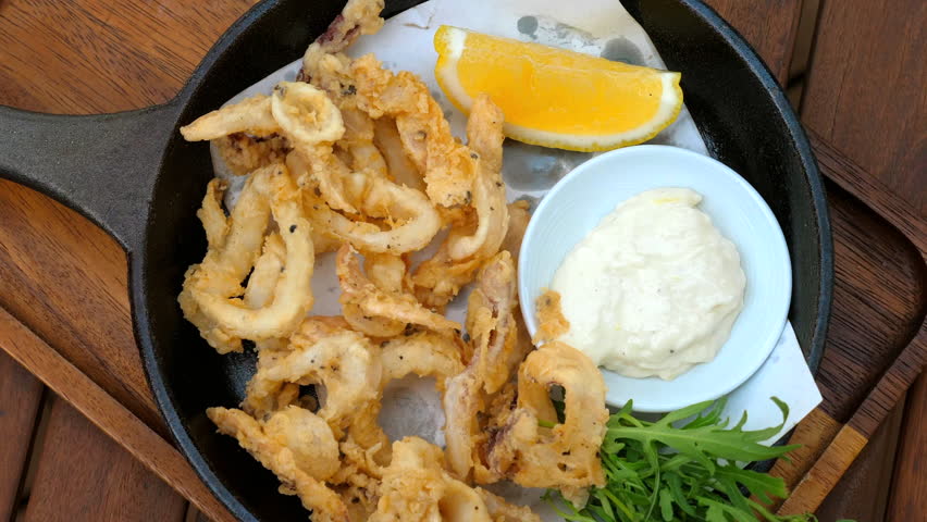 Hand dipping fried calamari into sauce with lemon wedge on black skillet, wooden table background.