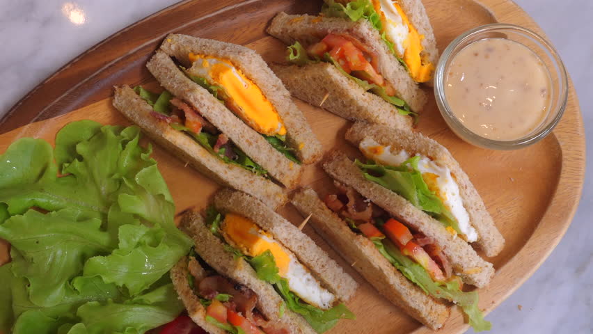 Delicious club sandwiches with eggs, bacon, lettuce, and tomato on whole wheat bread served on wooden platter, accompanied by fresh green lettuce and small dish of creamy dressing. Food and Cuisine.