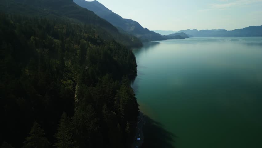 Drone flying around the lakes edge of Harrison Lake in British Columbia Canada on a hazy sunny morning with the large emerald lake water sitting very still and calm surrounded by hills of green trees