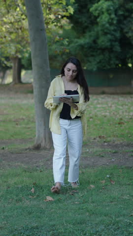 young brunette caucasian girl walking towards camera on the grass typing on the touch pad of a tablet