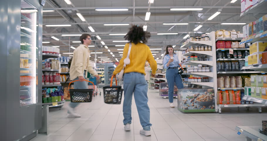Black Woman in a Yellow Sweater and Jeans Dancing in a Busy Supermarket, Carrying a Basket, Creating a Lively and Energetic Atmosphere. Customers are Shopping in the Background. Back View Footage