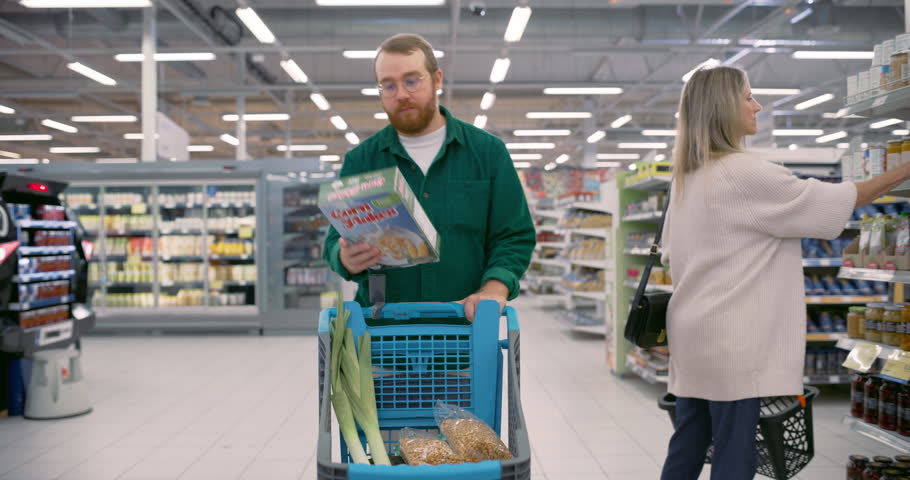 Male Shopper Walking with a Shopping Cart Between the Aisles of a Grocery Store. Cheerful Man Holding a Pack of Corn Flakes. Families, Friends, and Individuals Search for Fresh Food Produce