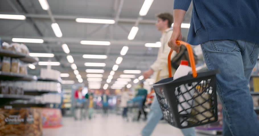 Anonymous Man Carrying a Shopping Basket Through a Modern Grocery Supermarket, Navigating Aisles Filled with Various Products and Other Shoppers. Low Angle Follow Footage