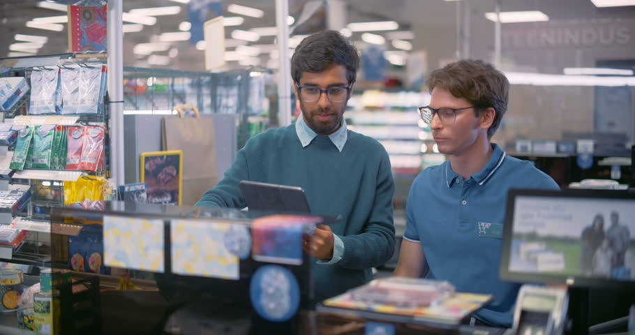Caucasian Male Supermarket Cashier in a Blue Polo Shirt Discussing Checkout Procedures with a Young Indian Manager, Using a Tablet Computer for Checking Schedule and Work Plans in a Grocery Store