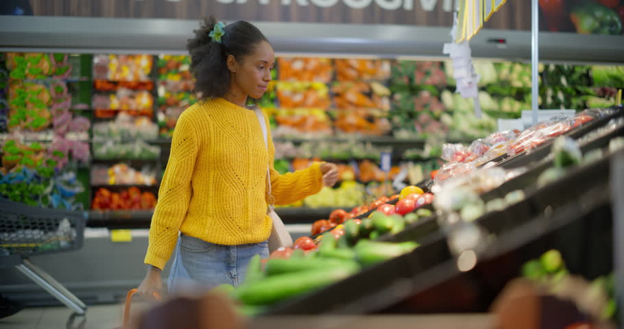 Stylish African American Woman in a Yellow Sweater Shopping for Fresh Produce in a Vibrant Supermarket Aisle, Carefully Selecting Cherry Tomatoes for Their Quality, Freshness and Price