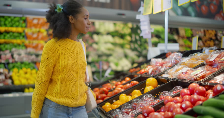 Beautiful Young Black Woman Shopping for Fresh Produce in a Modern Supermarket, Standing at a Vegetable Aisle and Carefully Selecting Tomatoes and Other Items From the Neatly Arranged Shelves