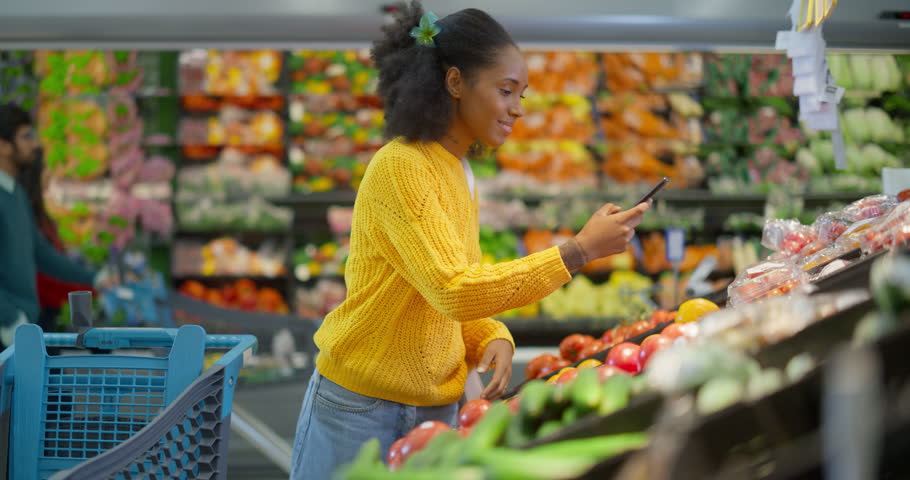 African Female Shopper Selecting Fresh Produce While Using Her Smartphone in a Supermarket, Using Device to Scan and Register Items Online as She Shops. She is Managing the List Through a Shopping App