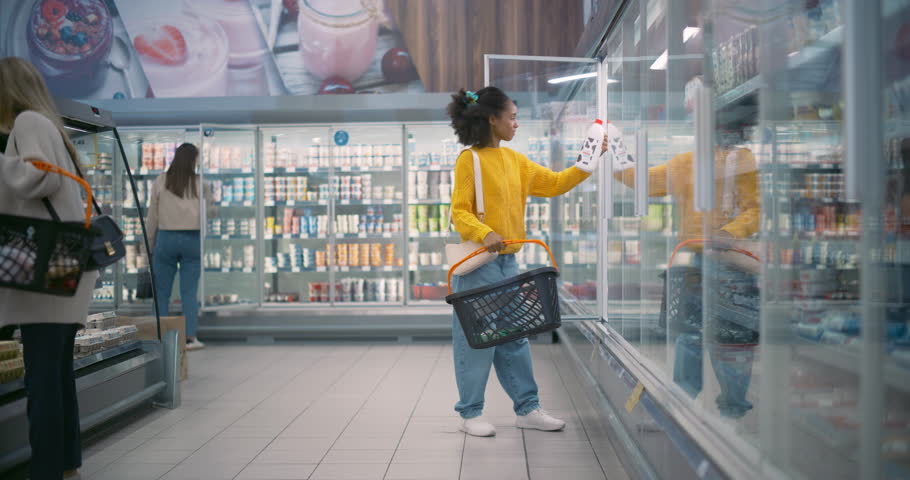 African Female Shopper Opens the Fridge Door in a Supermarket, Reaching for a Milk Bottle and a Dozen Eggs. She Places Them into Her Basket, Ensuring She Gets Fresh Ingredients for Dinner
