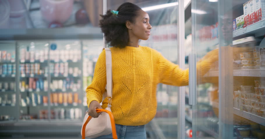 Young Black Woman Standing in Front of the Refrigerated Section, Opening the Fridge to Grab Milk and Eggs. She Quickly Adds Them to Her Basket, Planning Her Next Grocery Choices