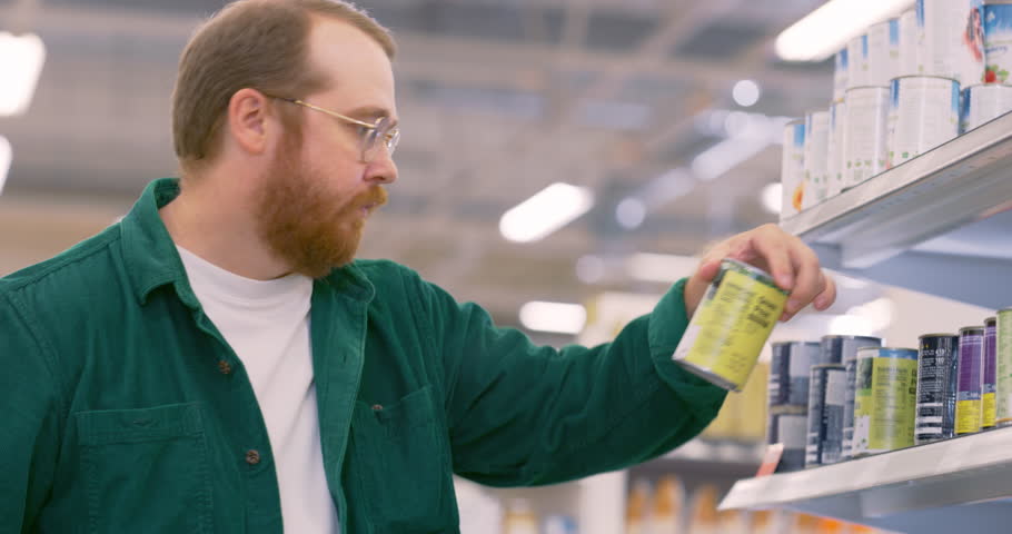 Male Customer Shops in a Busy Supermarket. Young Caucasian Man Carefully Selecting Healthy Groceries for Dinner. He is Choosing the Best Can of Green Peas from a Stocked Shelf with Various Items