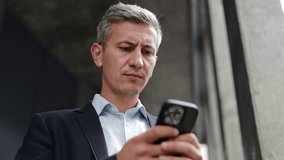 A businessman in a suit, standing outdoors, frowns and looks concerned as he reads a message or email on his phone. This conveys negative business news, disappointment, stress, and uncertainty. - Powered by Shutterstock - Get 15% off with code: PIKWIZARD15