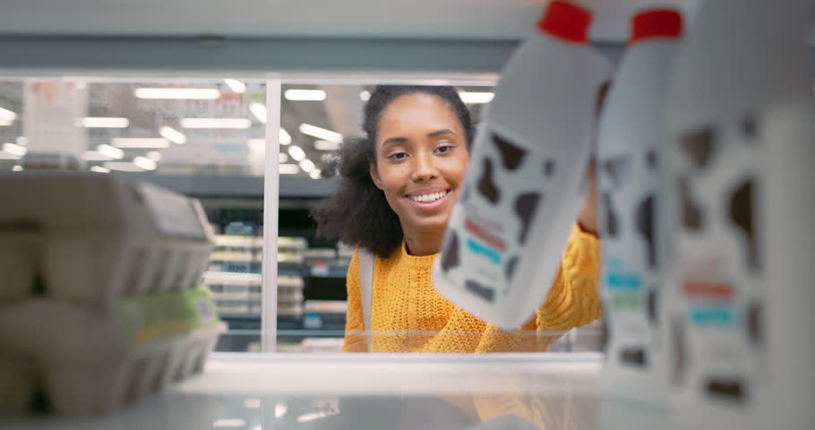 Beautiful Young African American Woman in a Yellow Sweater Takes a Milk Bottle and a Pack of Eggs From a Supermarket Fridge. POV Portrait of a Grocery Shopper From Inside the Fridge