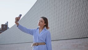Beautiful brunette taking selfie on smartphone near modern town architecture closeup. Confident woman in stylish blue dress capturing joyful moment in urban street. Cheerful female blogger using phone - Powered by Shutterstock - Get 15% off with code: PIKWIZARD15