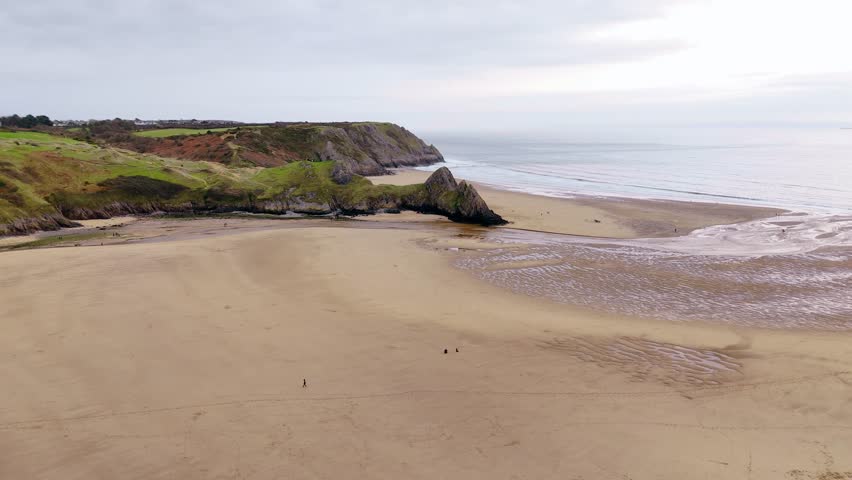 People walking across the vast sandy beach of Three Cliffs Bay during low tide