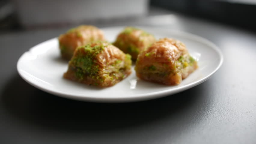 Four Greek or Turkish baklava pieces on a white plate, each topped with crushed pistachios. The layers of golden, flaky pastry glisten, highlighting the rich, sweet desert filling and inviting texture